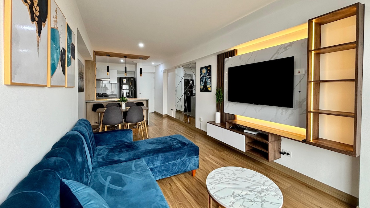 A modern living area features a spacious sectional sofa in a deep blue hue, paired with a marble coffee table. A large wall-mounted TV is framed by a combination of cabinetry and decorative art. The open layout connects seamlessly to a well-equipped kitchen in the background.