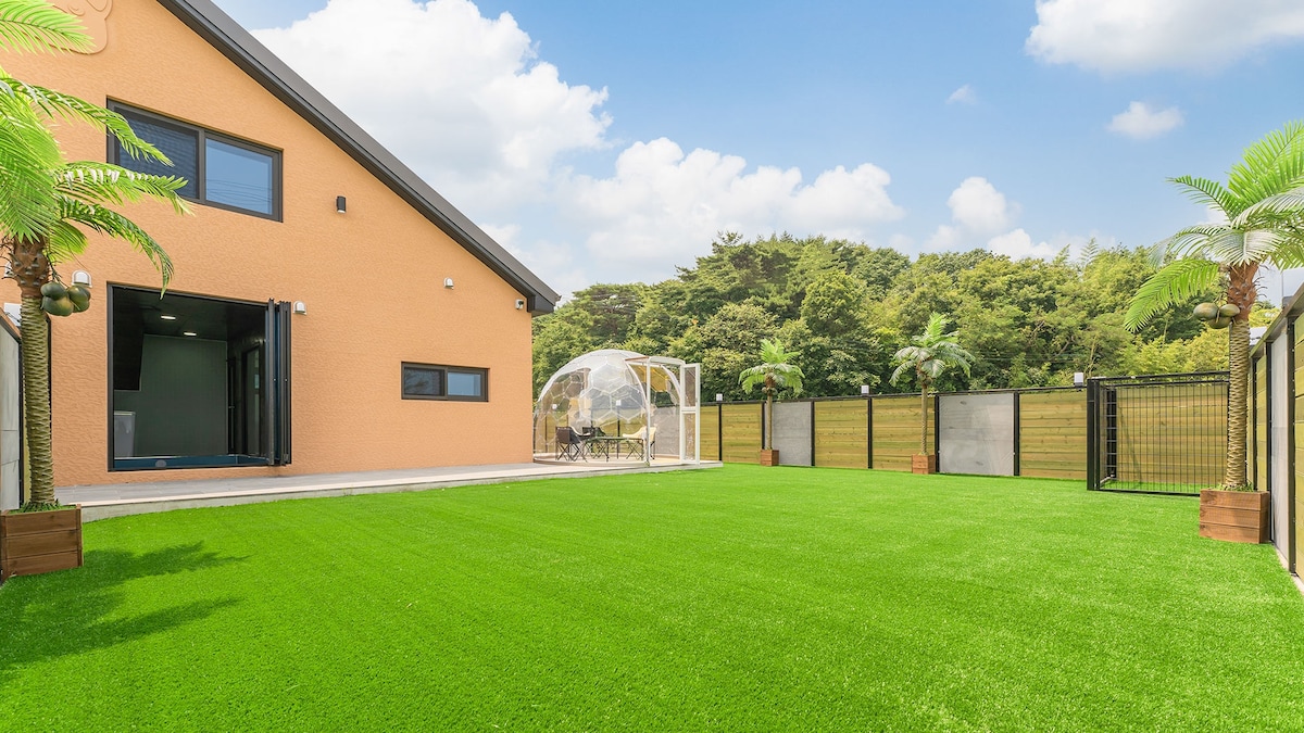 An outdoor area showcases a spacious, well-maintained lawn framed by greenery. A transparent dome for dining is visible in the distance, alongside modern fencing that provides privacy. The building features large windows and a welcoming entrance, enhancing the overall inviting atmosphere.