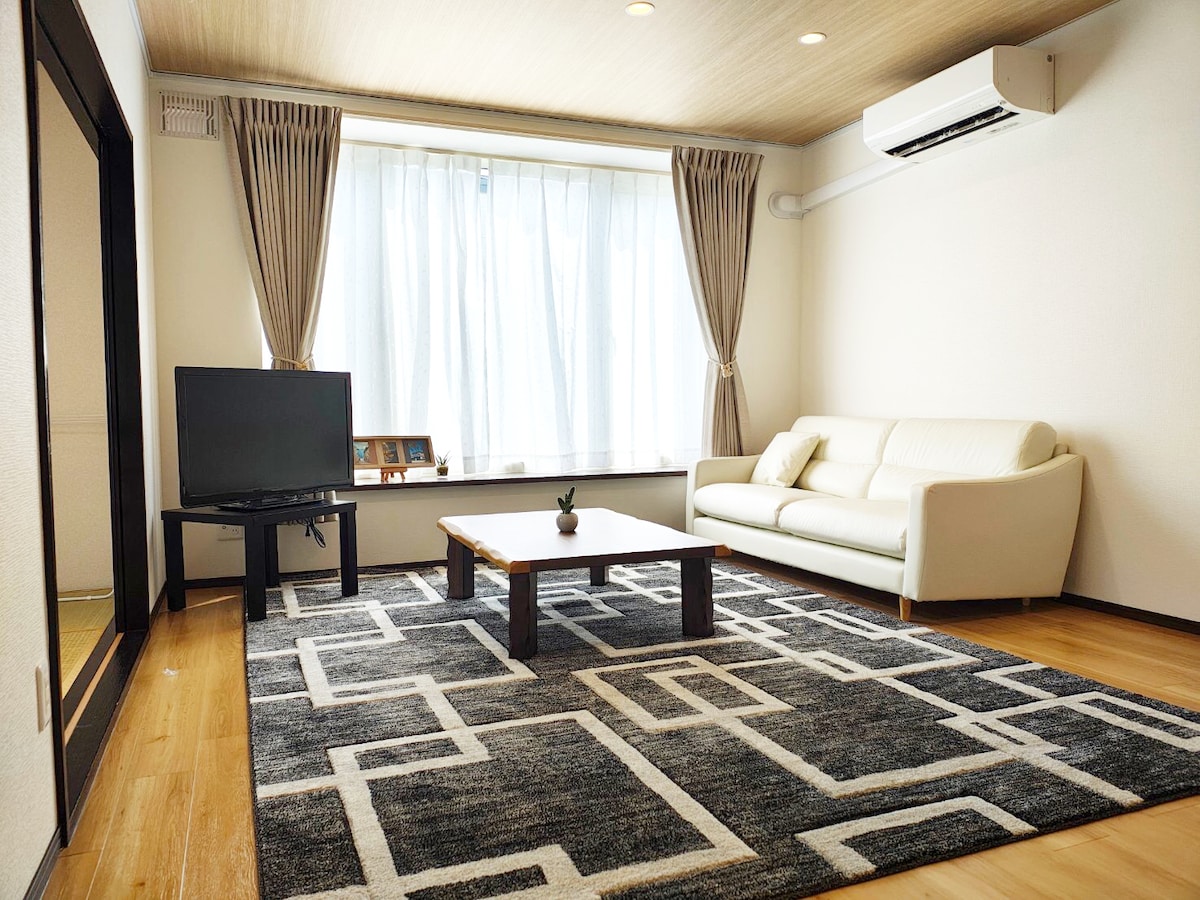 The living area is illuminated by natural light flowing through large windows with soft drapes. A comfortable white sofa and a low wooden table sit on a textured rug, complemented by a television nearby. A potted plant adds a touch of greenery to the space.