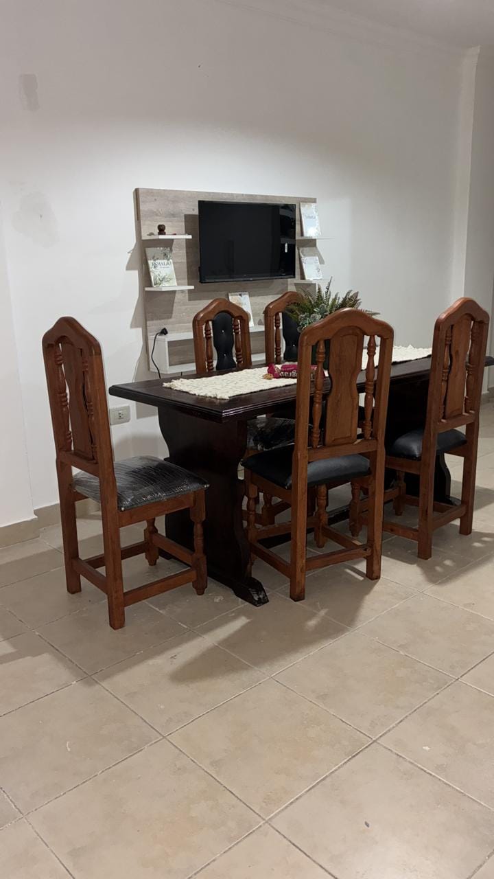 A wooden dining table is surrounded by four high-backed chairs. A simple decorative piece is placed on the table's center. A wall-mounted TV is visible above the table, accompanied by a minimalist shelf displaying small items.