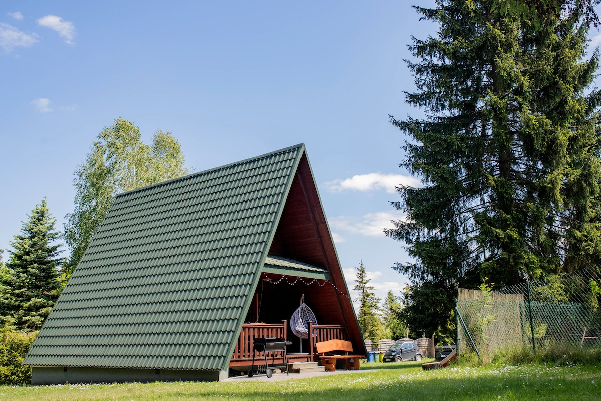 A charming cabin features a triangular roof covered in green tiles, set against a clear blue sky. Surrounded by lush grass and tall trees, a cozy porch with wooden seating creates an inviting entryway.