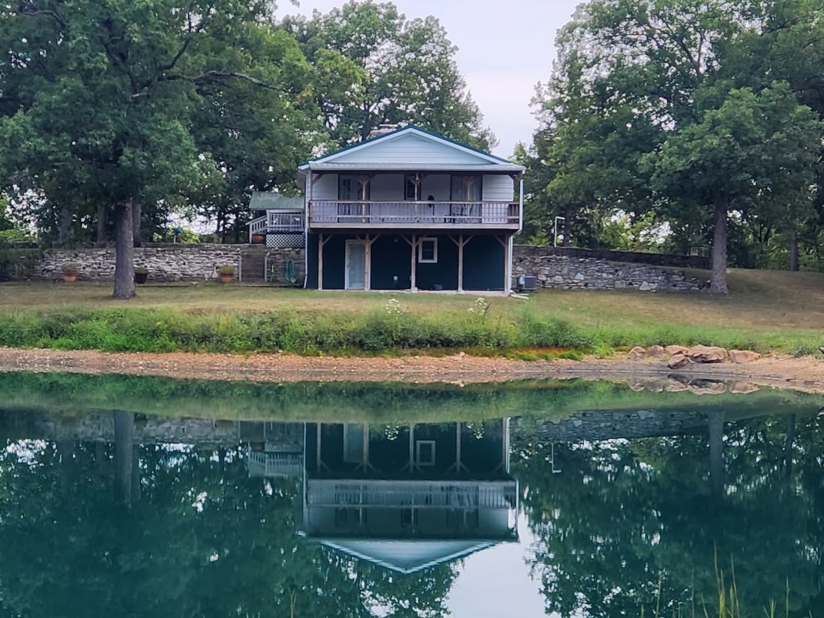 A cozy farmhouse is situated on nine acres, surrounded by mature trees. The building is reflected in the calm water of a pond, showcasing its two-story structure with a covered porch on the upper level, framed by greenery.