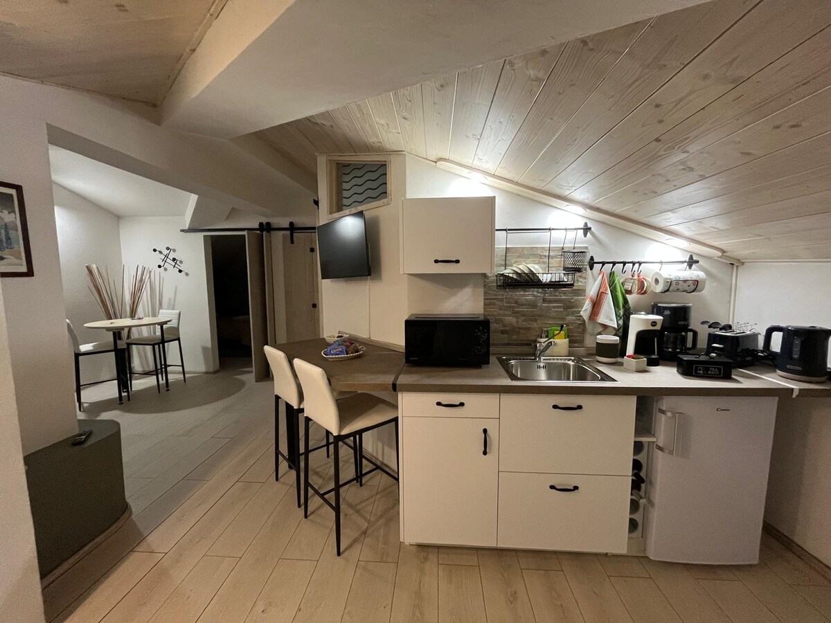 An open kitchen area features modern appliances and a wooden countertop. Two high stools are positioned at the counter, while a dining table with chairs is visible in the background. The ceiling is angled, enhancing the cozy atmosphere of the space.