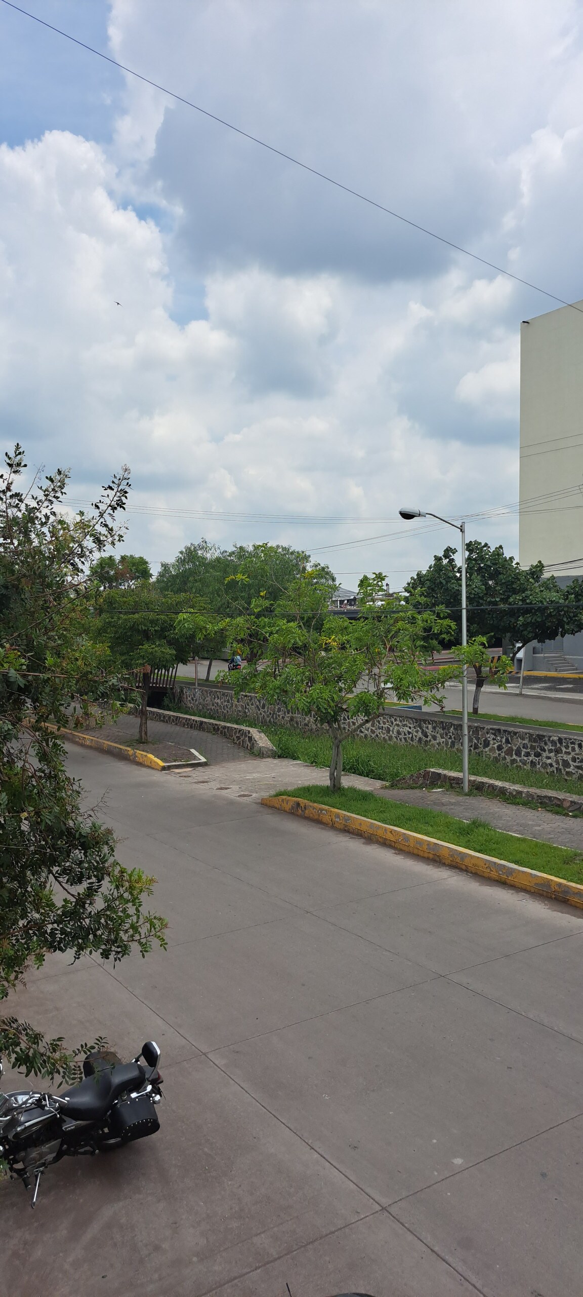 A view of a tree-lined street is presented, featuring a smooth asphalt surface. Various greenery is visible along the roadside, with scattered clouds in the sky above. A motorcycle is parked near the curb, and light poles are positioned along the edge of the road.