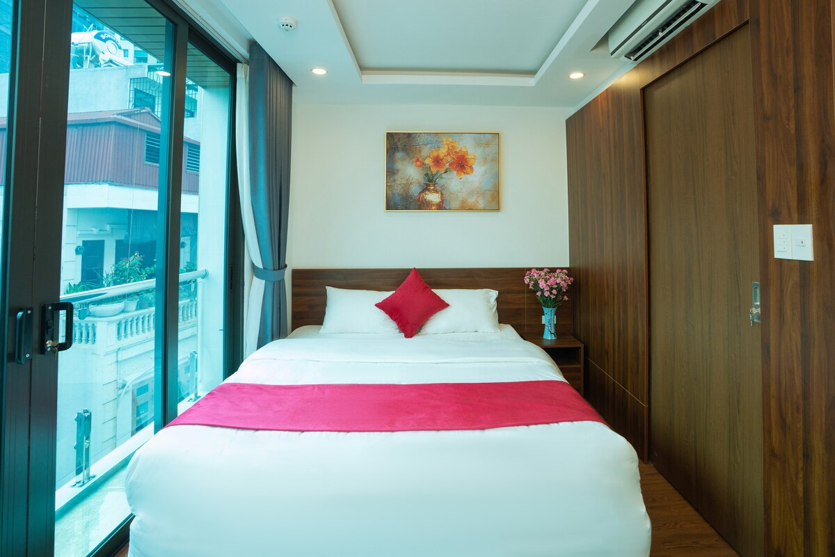 A well-furnished bedroom features a spacious bed adorned with a white duvet and a pink accent blanket. A decorative wall painting enhances the space, and fresh flowers are displayed in a vase on a nightstand. Large windows provide natural light and views of the neighborhood.