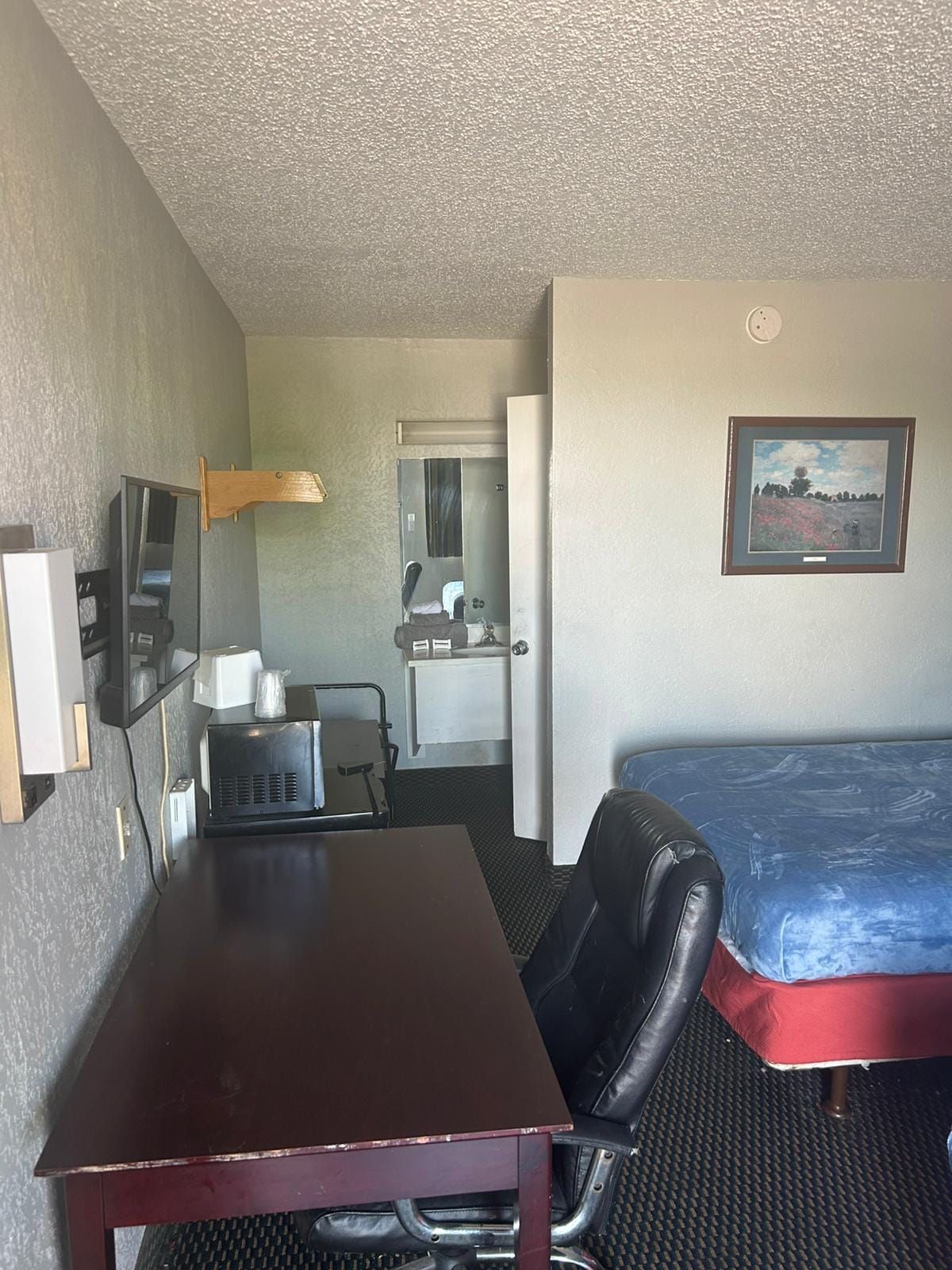 The room features a spacious bed positioned beside a functional desk and chair. A wall-mounted television is visible, along with a mini fridge and coffee maker on a shelf. A door leads to a separate area, while natural light enters through a window.