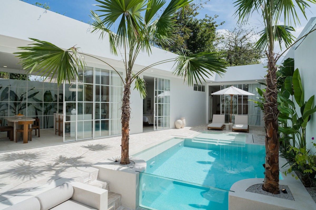 A serene outdoor space features a private pool surrounded by tropical palm trees. Sun loungers are positioned beside the pool, while an umbrella provides shade. Expansive glass doors lead to the enclosed living area, enhancing the connection between indoor and outdoor environments.