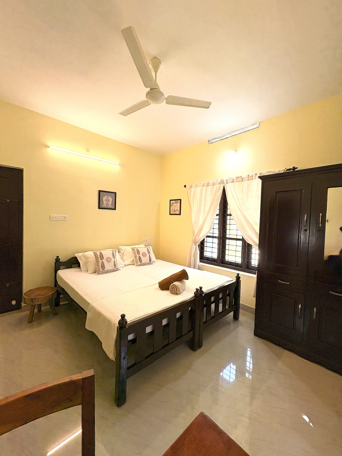 A well-lit bedroom features a spacious bed with an off-white bedspread, complemented by decorative pillows. A wooden stool is positioned beside the bed. Two dark wood wardrobes are visible, and light flows through a window dressed with sheer curtains, illuminating the yellow walls.