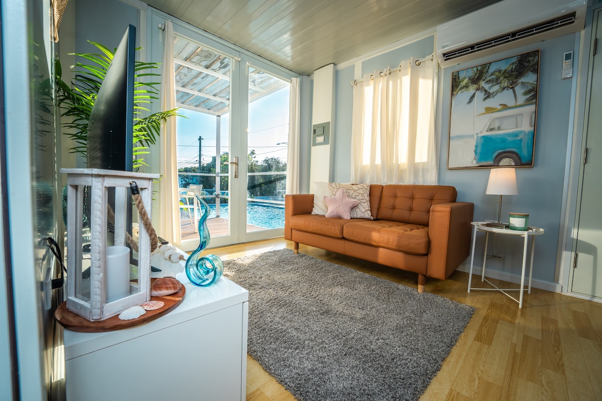 A light-filled living area features a brown sofa facing large glass doors that open to a private outdoor space. A soft area rug lies beneath a small coffee table, with decorative items on display. Light curtains frame the windows, enhancing the bright ambiance.