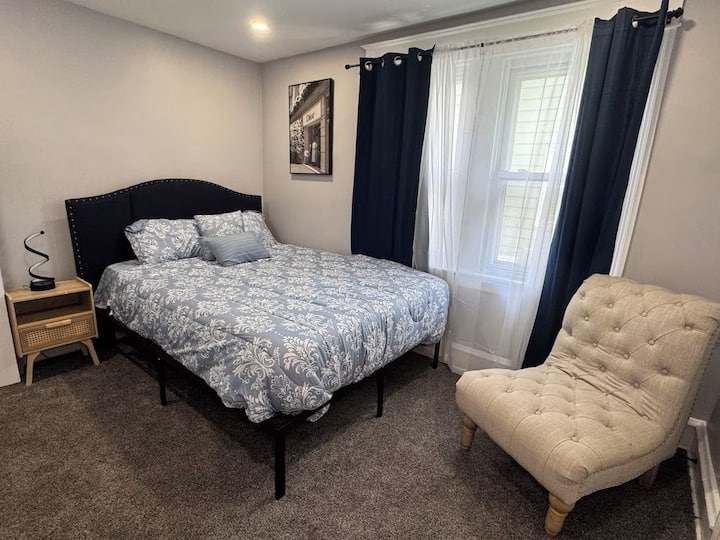 Útulná spálňa s pohodlnou manželskou posteľou, nástennou televíziou, plyšovým kreslom a prirodzeným svetlom z okna s tmavými závesmi pre súkromie.