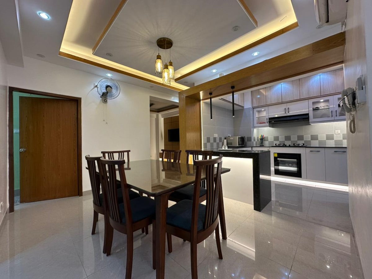 A modern dining area features a large dark wood table surrounded by six matching chairs. An open kitchen design is visible in the background, displaying sleek cabinetry and stainless steel appliances. Ambient lighting is provided by a stylish ceiling fixture, enhancing the contemporary feel.