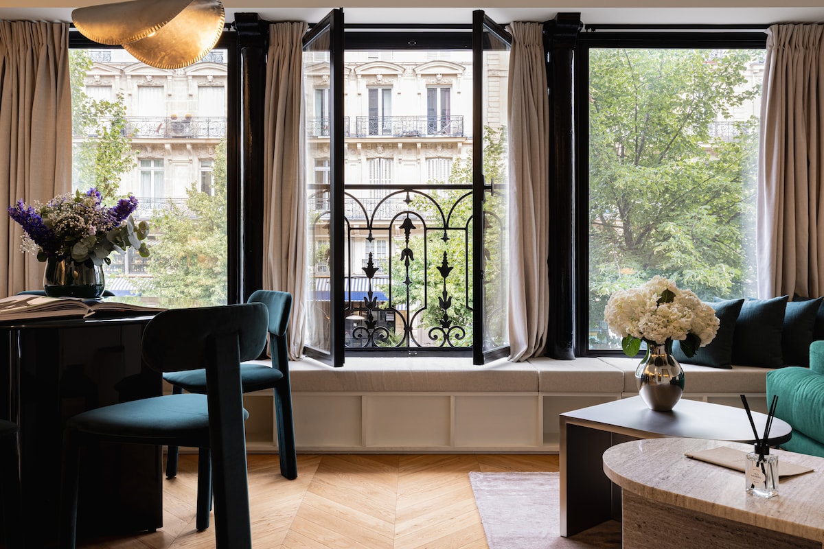 A bright living space features large windows with black wrought iron detailing, allowing views of greenery outside. The seating area includes a cushioned bench and a round table with dark chairs. Fresh flowers in a vase add a touch of nature to the room.