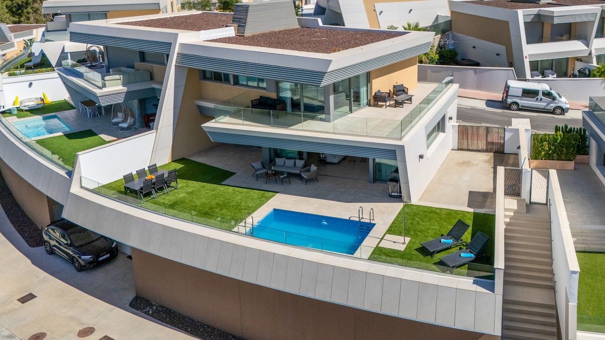 An aerial view of a modern villa showcases multiple terraces and a private pool framed by neatly trimmed grass. Spacious seating areas are arranged on the balconies, with sun loungers positioned beside the pool. The sleek architectural lines and natural materials create a contemporary aesthetic.