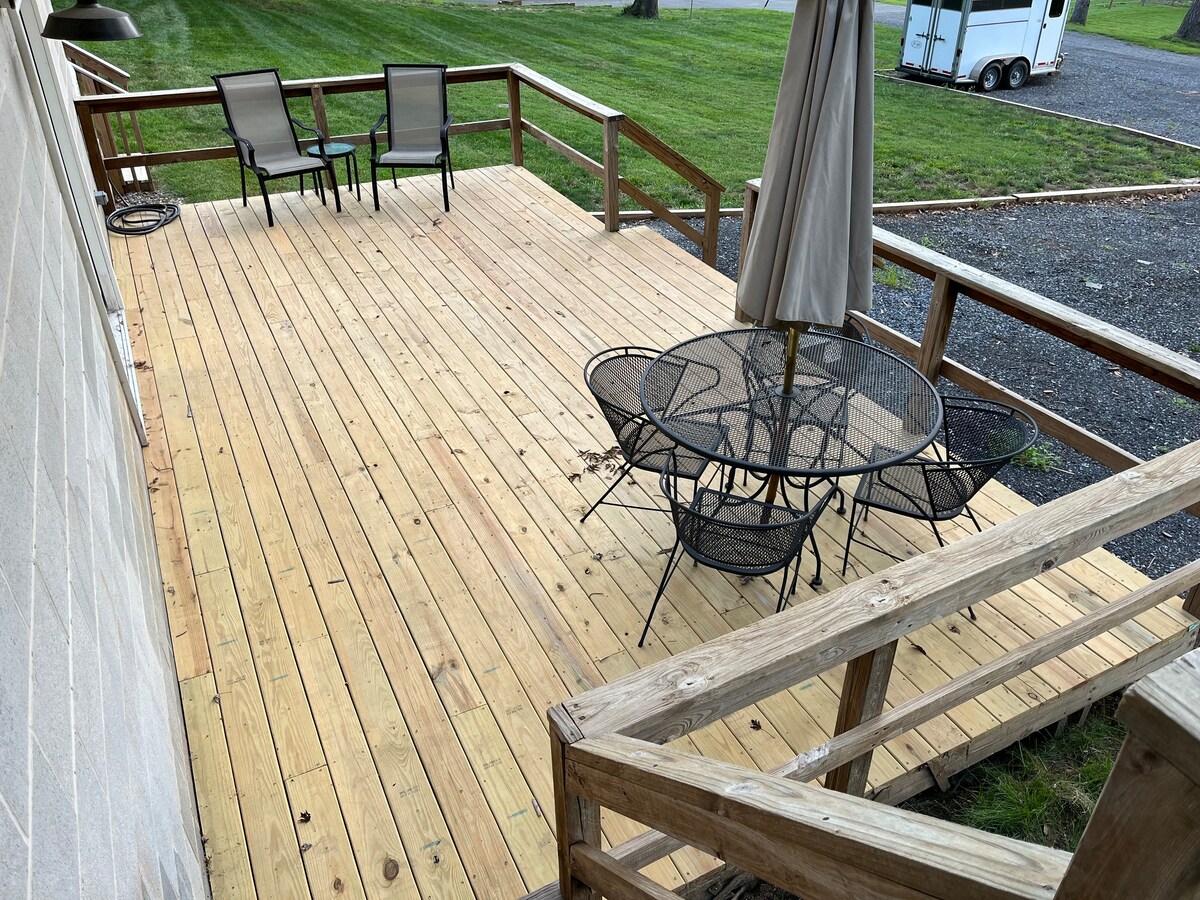 An outdoor deck is shown, featuring a circular table surrounded by four black wire chairs. Two additional lounge chairs are positioned nearby. A large umbrella is positioned at the table, offering shade. The deck overlooks a well-maintained grassy area.