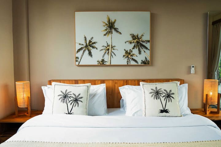 Genieße erholsame Nächte in diesem Zimmer im tropischen Stil mit weichen Kissen, Kunst mit Palmenmotiven und sanfter Beleuchtung – dein privater Rückzugsort auf der Dachterrasse der Villa Yaro.
