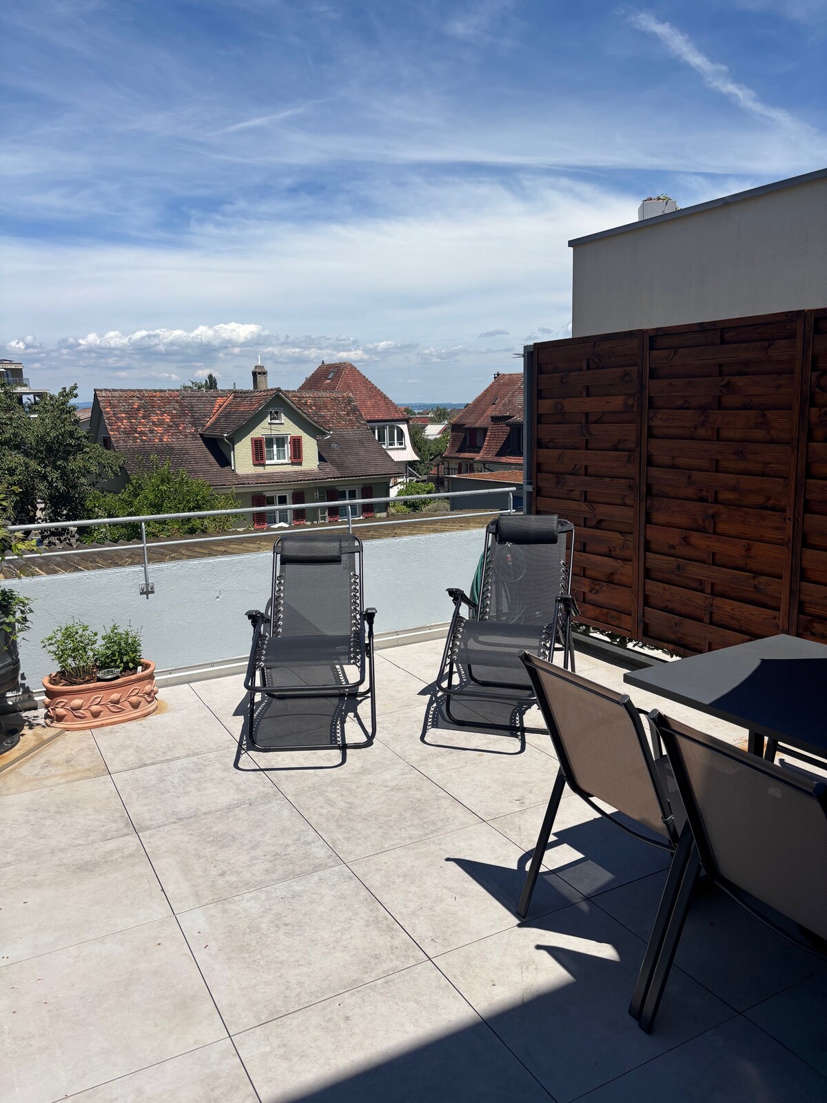 A terrace features outdoor seating with two reclining lounge chairs and a dining table. The area is surrounded by a low wooden screen and offers views of nearby houses and the distant landscape under a clear sky.