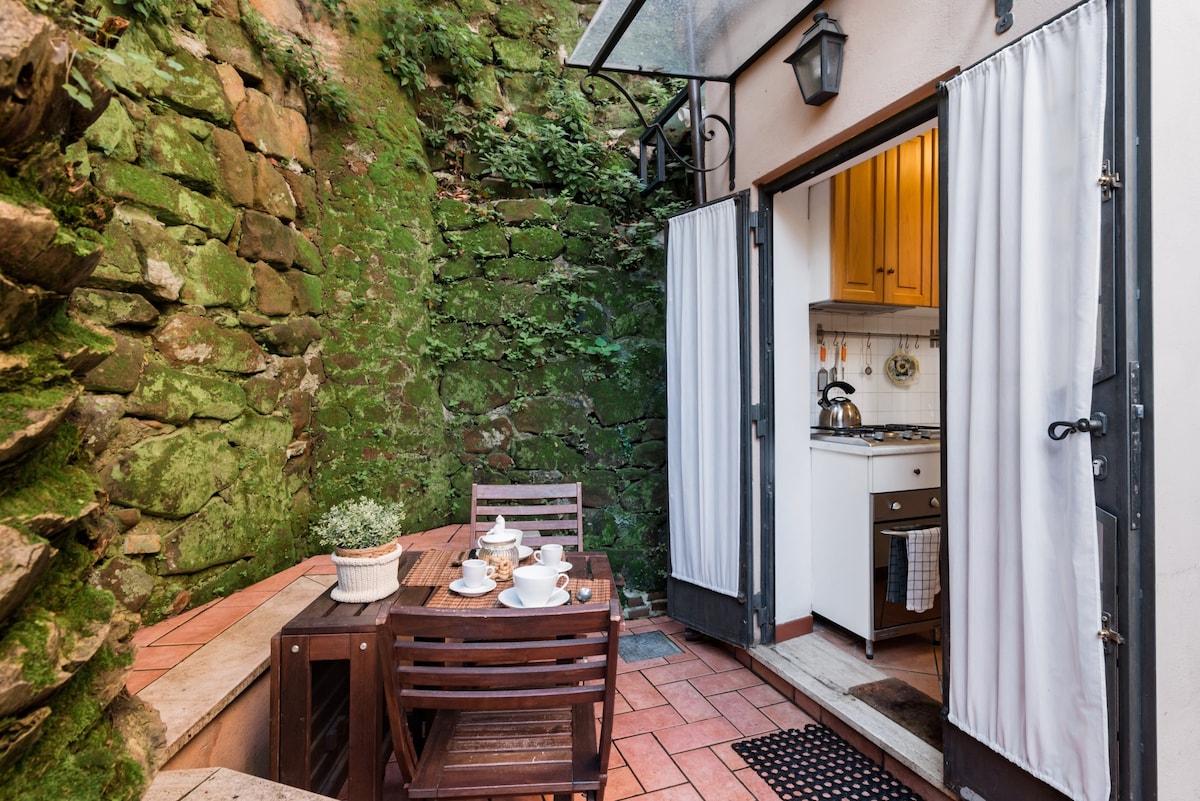 A charming outdoor patio is framed by a natural stone wall, creating a tranquil space. A wooden table is set with teacups, while a small potted plant adds a touch of greenery. The entrance to the kitchen is visible, with light wooden cabinetry and tiles hinting at a cozy atmosphere.