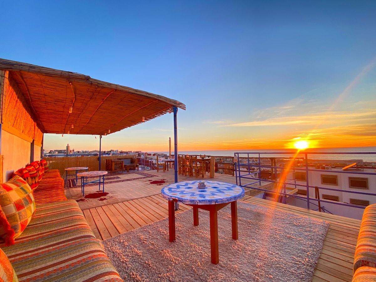 A spacious terrace offers comfortable seating arranged around a patterned table, with colorful cushions adding warmth. The sun sets in the distance, casting a golden light over the surrounding rooftops and creating a serene outdoor space for relaxation.