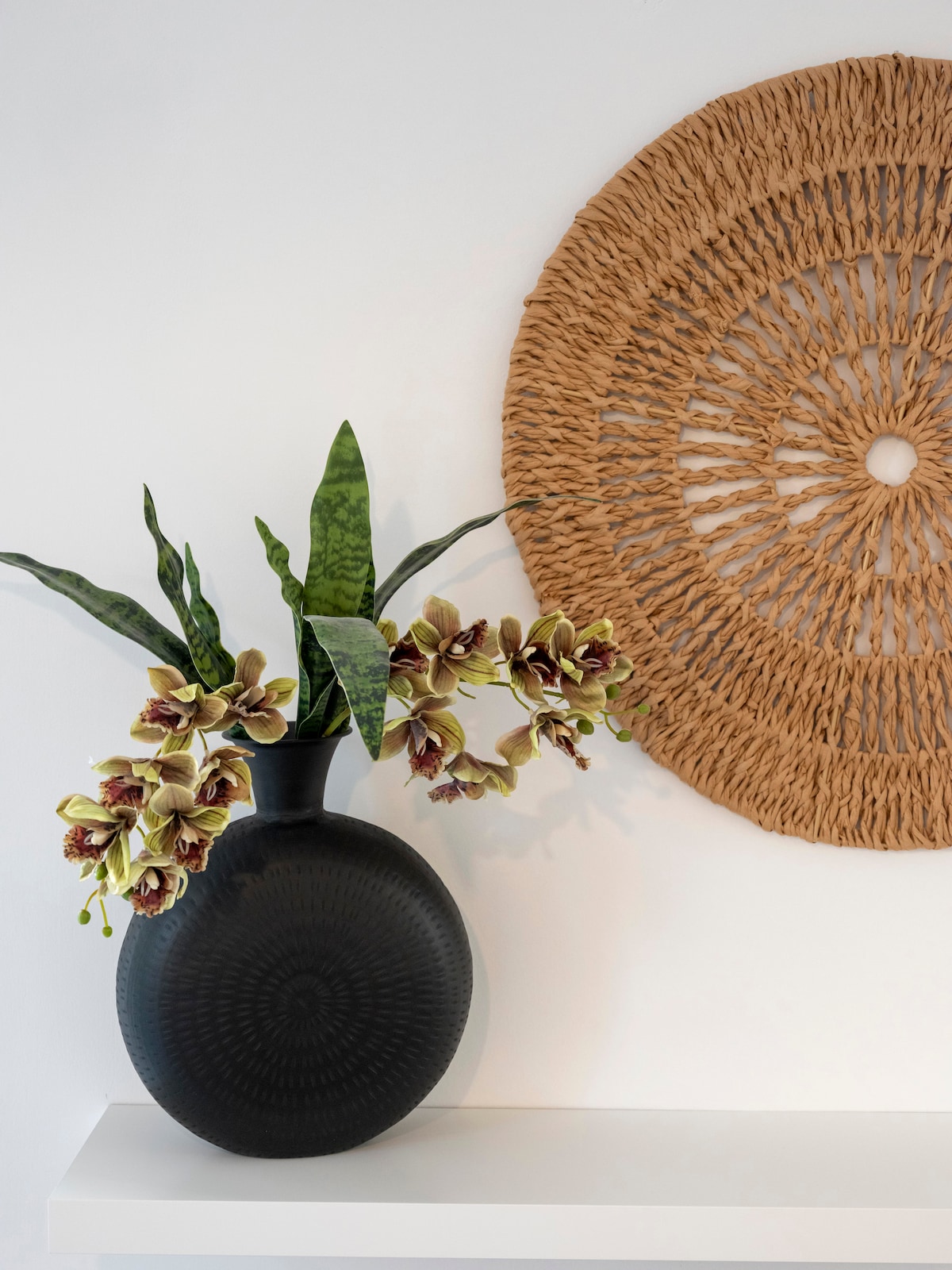 A decorative black vase is positioned against a light wall, adorned with a cluster of artificial flowers in soft green and cream hues. To the right, a circular woven wall hanging adds texture and warmth to the space.
