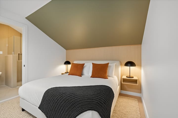Cette chambre dispose d'un très grand lit, d'une salle de bain privée avec baignoire et douche, d'un plafond gris en pente, d'un éclairage chaleureux et de tables de chevet flottantes avec des lampes noires modernes.