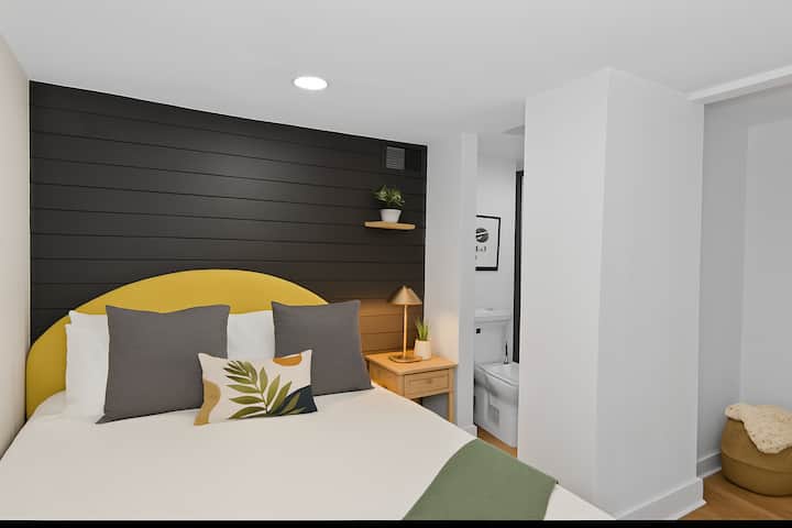 Cette chambre confortable au niveau inférieur dispose d'un grand lit avec une tête de lit jaune et d'une décoration moderne. Il comprend une salle de bain privée avec douche complète et dispose de sa propre entrée sans marches — accès facile et pratique.