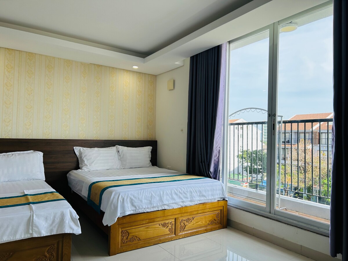 A well-lit guest room features two spacious beds with white linens and decorative blue accents. Large windows offer views of the outdoors, while soft curtains frame the glass, enhancing natural light within the space. A modern, minimalistic design is evident throughout the room.