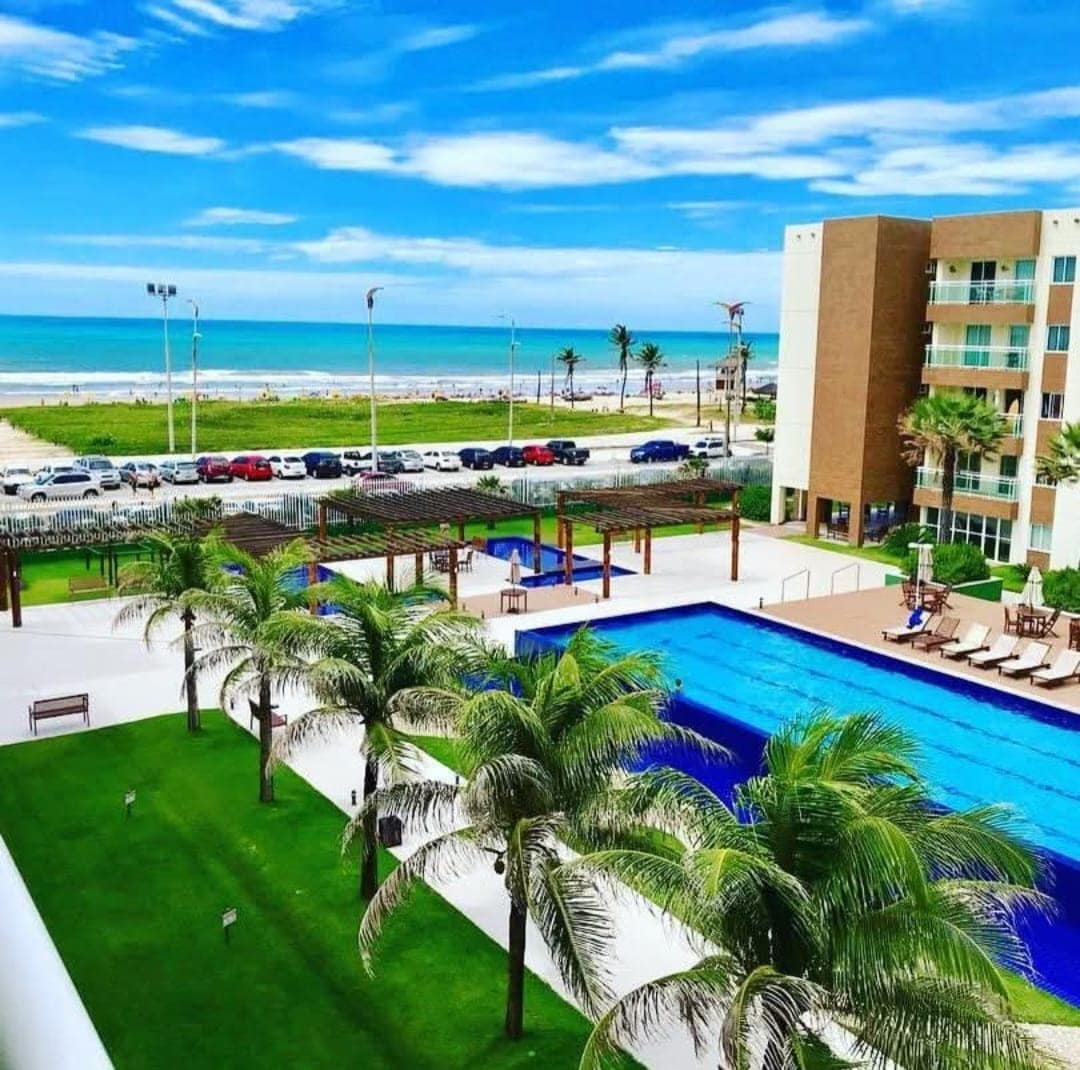 An aerial view captures a spacious pool surrounded by lush green grass and palm trees, providing a refreshing outdoor leisure area. The beach is visible in the distance, complemented by clear blue skies. The nearby building showcases multiple stories and expansive windows.