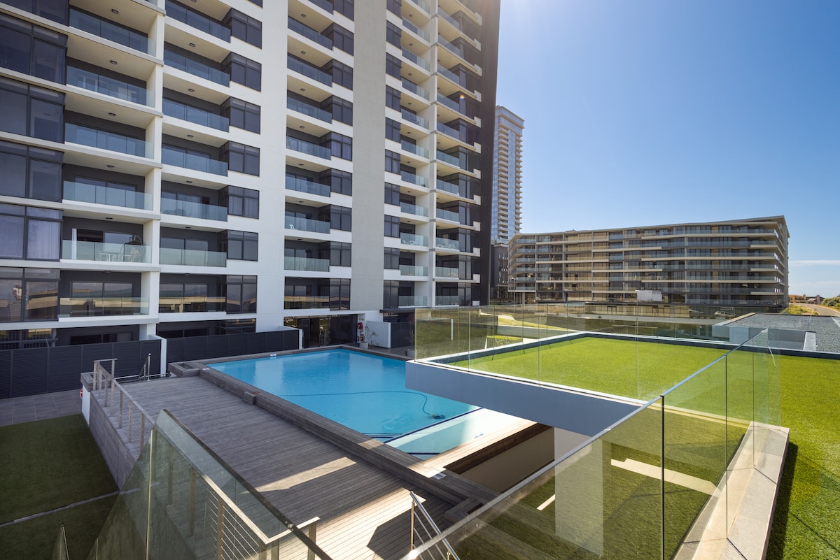 The image showcases a modern apartment building with a well-maintained outdoor space. A large swimming pool is bordered by a wooden deck and grassy area, surrounded by glass fencing. The structure's sleek facade reflects the clear blue sky.