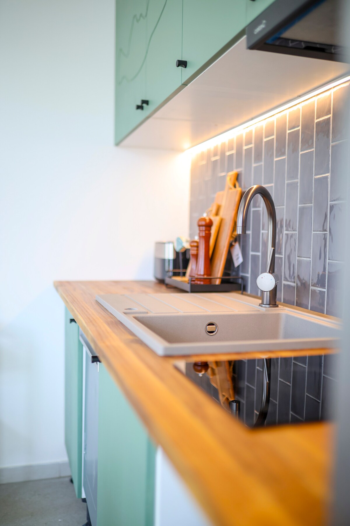 A modern kitchen showcases sleek cabinetry in light green, complemented by a wooden countertop. A minimalist sink with a modern faucet is positioned against a stylish tiled backsplash, adding to the functional design. Various kitchen utensils can be seen on the countertop.