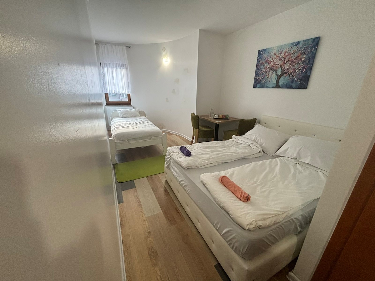 A shared sleeping area contains two beds, one spacious and one smaller. Light-colored linens are neatly arranged on both beds, and a small table with green chairs is positioned in the corner. Natural light enters through a nearby window, brightening the room.