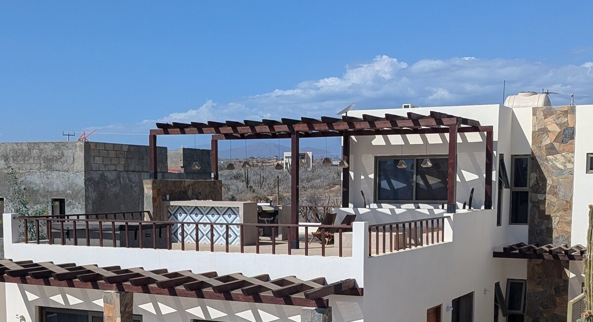 The image showcases a rooftop terrace with a pergola structure, providing shade over a spacious outdoor area. The surroundings are characterized by a clear blue sky and distant mountains, with natural stone and white walls adorning the building's facade.
