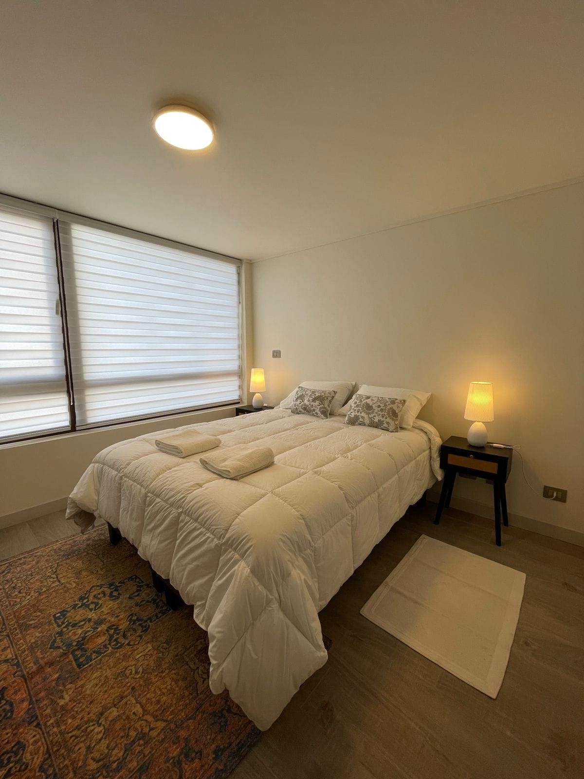 A well-lit bedroom features a spacious bed dressed in crisp white linens, complemented by decorative pillows. Soft lighting from bedside tables illuminates the area, while large windows with layered shades allow natural light to softly filter in, enhancing the room's calm atmosphere.
