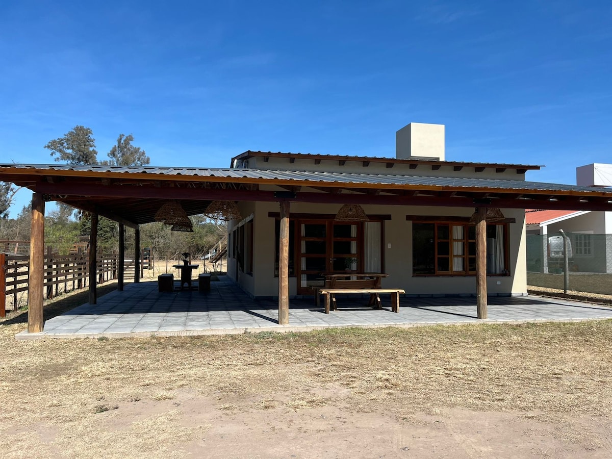 A single-story house is showcased, featuring a spacious covered patio with wooden beams and two hanging lights. A large wooden table is placed on the tiled patio, surrounded by ample outdoor space. The exterior is complemented by a clear blue sky and surrounding greenery.