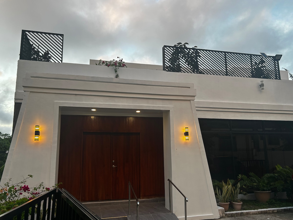 The exterior of Villa Sóller features a modern, minimalist facade with warm wooden doors and accent lights. A staircase leads up to the entrance, flanked by greenery, and a rooftop area is visible above, showcasing railing and potted plants against a cloudy sky.