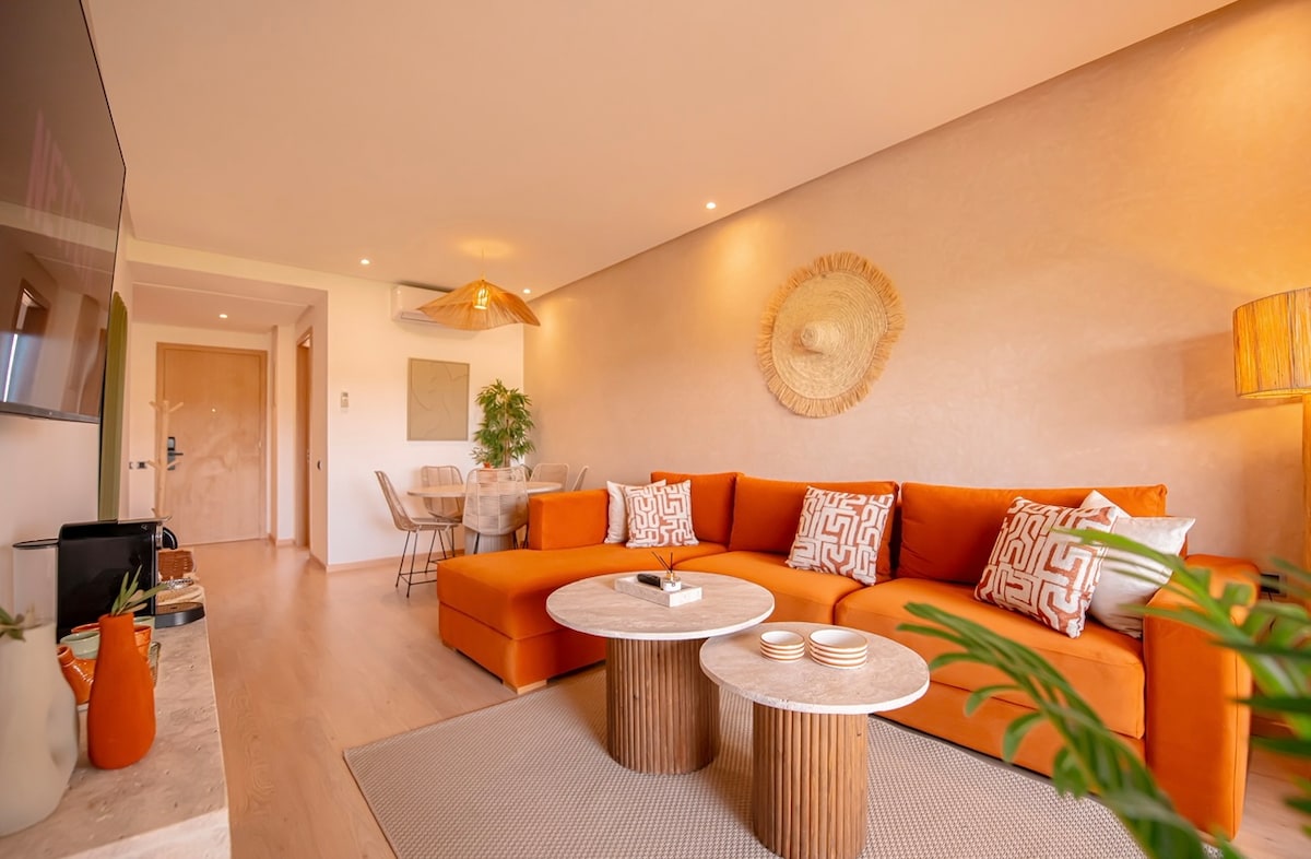 A bright living area is highlighted by a spacious orange sectional sofa adorned with patterned throw pillows. Natural light filters in, showcasing textured accent walls and contemporary decor. Two round coffee tables sit on a soft area rug, complemented by a dining table positioned in the background.