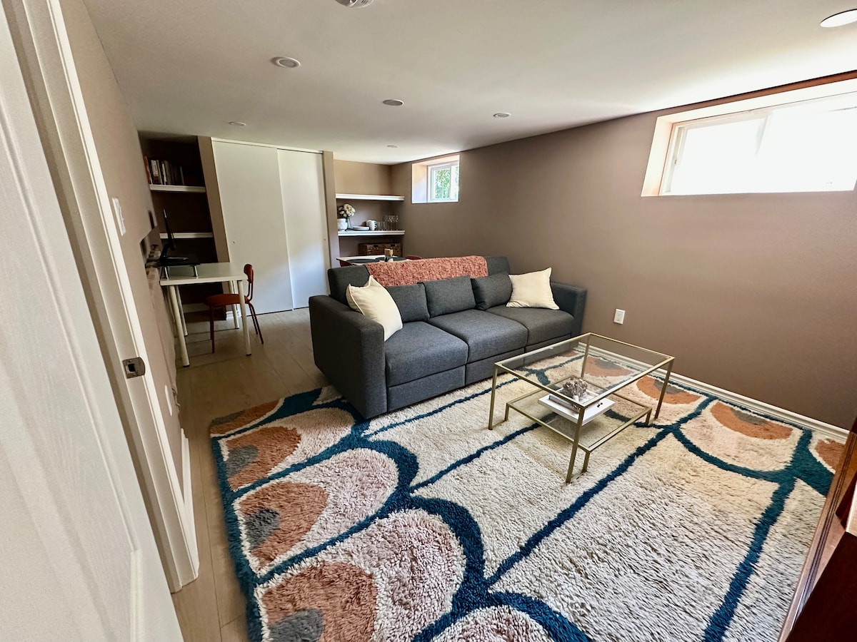 A cozy living room is featured, highlighted by a spacious gray sofa adorned with decorative pillows. A glass coffee table sits atop a patterned rug, while natural light enters through a small window. A desk with a chair is positioned against the wall, providing a designated workspace.