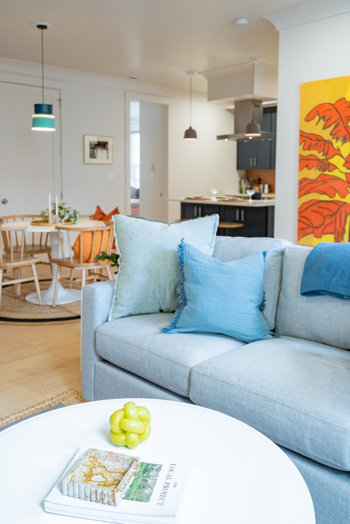 A light-filled living area features a gray sofa adorned with textured cushions in blue hues. A round coffee table holds a decorative plate with fruit. In the background, a dining space is visible, accompanied by a modern kitchen with dark cabinetry and pendant lighting.