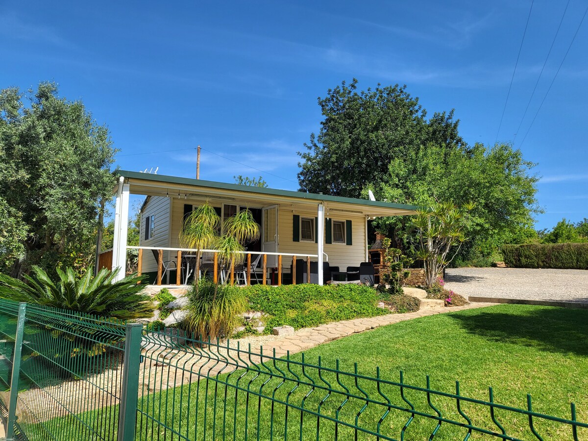 A charming bungalow is set within a landscaped garden, featuring a front porch shaded by a roof. Lush greenery and decorative plants surround the pathways, while a gravel parking area is visible in the background, enhancing the welcoming outdoor space.