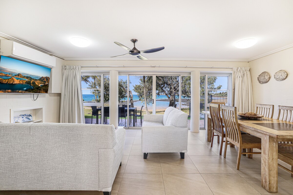 The spacious lounge area features large glass doors that open to stunning water views. A comfortable white sofa and a dining table are visible. Natural light illuminates the room, enhancing the serene atmosphere. Decorative artwork is displayed on the walls.