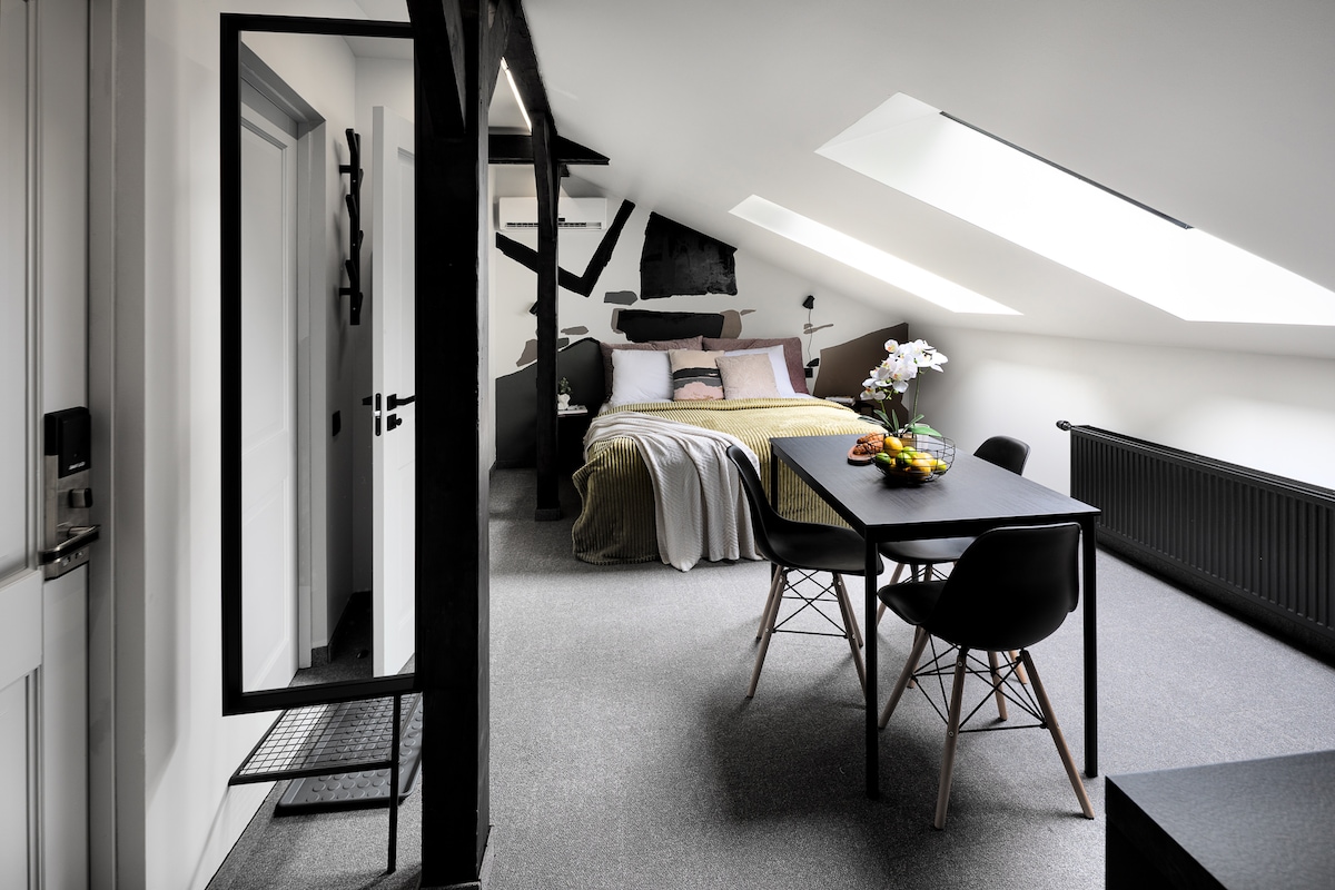 A bright loft apartment features a spacious sleeping area with a large bed and decorative pillows. A dining table with black chairs is positioned beside a window, complemented by a fruit arrangement. Skylights illuminate the room, enhancing its airy feel.