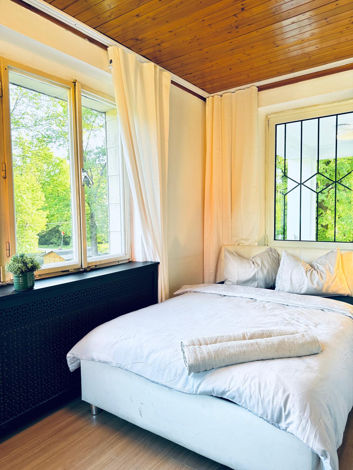 A serene bedroom features a spacious bed with layered white bedding and decorative pillows. Large windows allow natural light to brighten the space, framed by soft curtains. A minimalistic bedside table adds functional charm, while greenery is displayed on the window ledge, enhancing the room's tranquil ambiance.