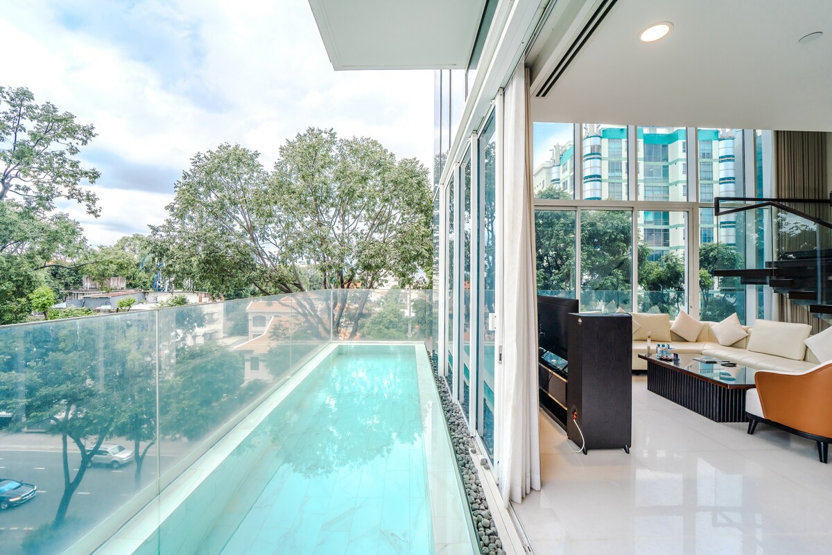 A bright outdoor terrace features a clear glass railing overlooking a tranquil pool. The adjoining indoor area is filled with natural light, showcasing comfortable seating arranged around a low coffee table against a backdrop of large windows.