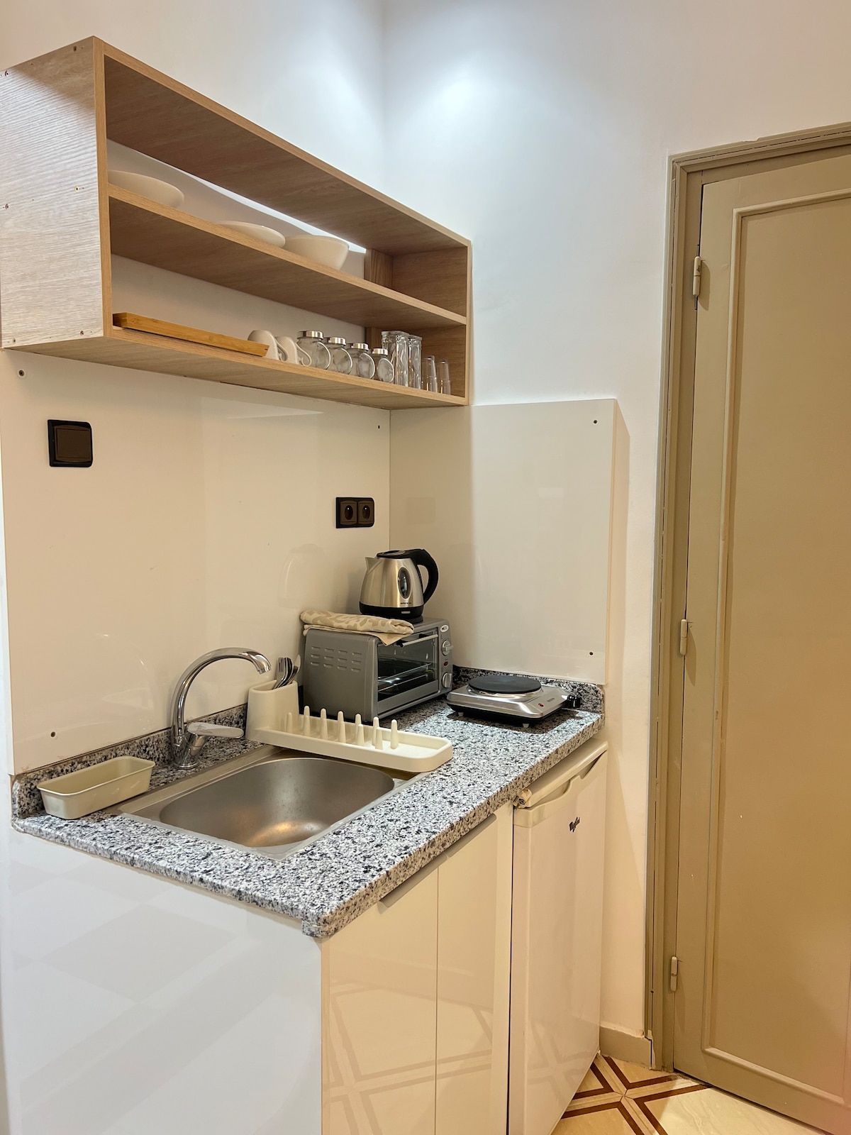 A compact kitchenette is presented with a stainless-steel sink and modern countertop. Basic appliances, including a kettle and a toaster, are neatly arranged. Glassware is stored on an upper shelf, contributing to a functional space. A door leads to an adjacent area.