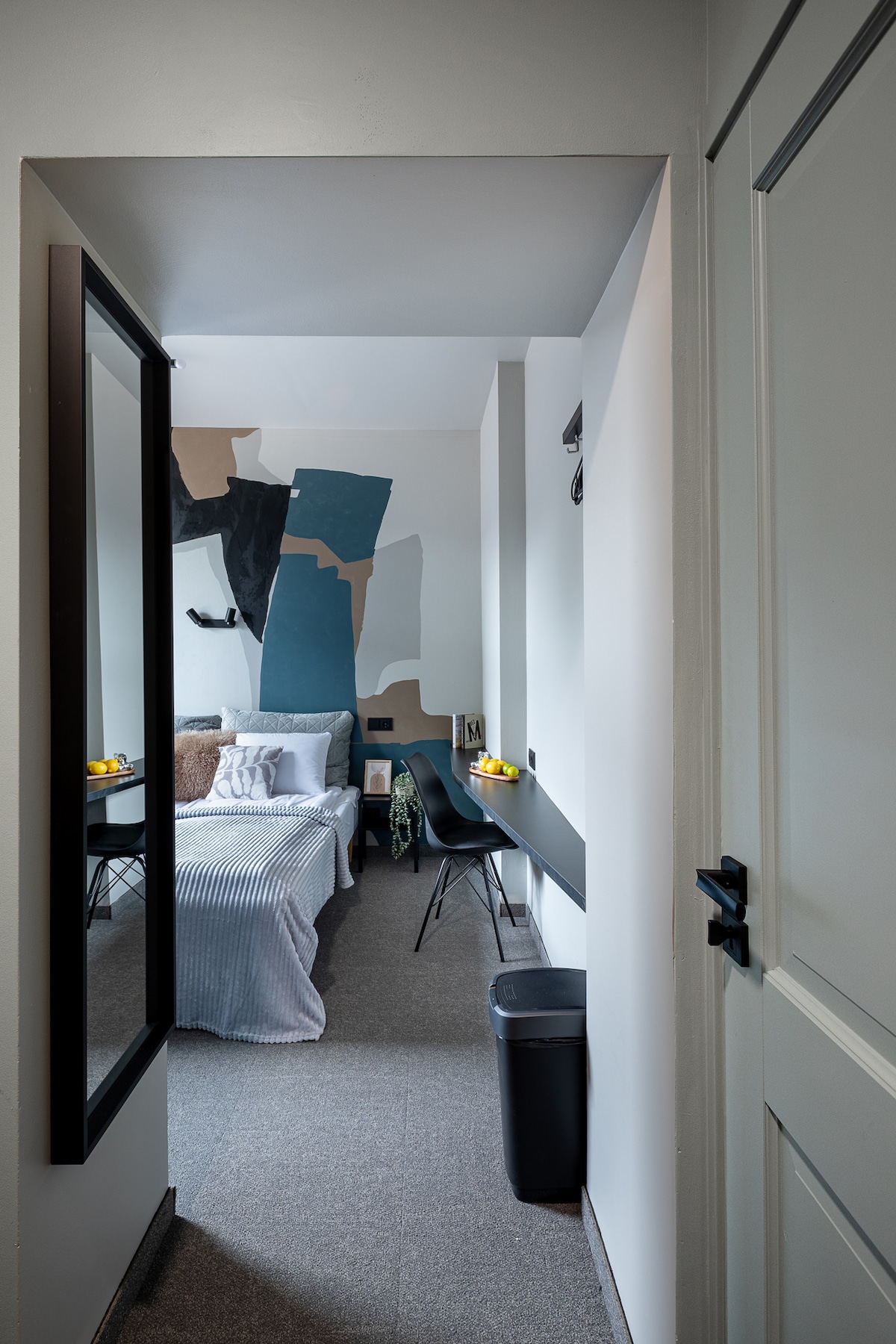 A view into a compact room reveals a comfortable sleeping area featuring layered bedding and a wall adorned with modern art. Natural light filters in through the space, highlighting a small workspace with a chair, while a mirror offers reflections of the room's design.