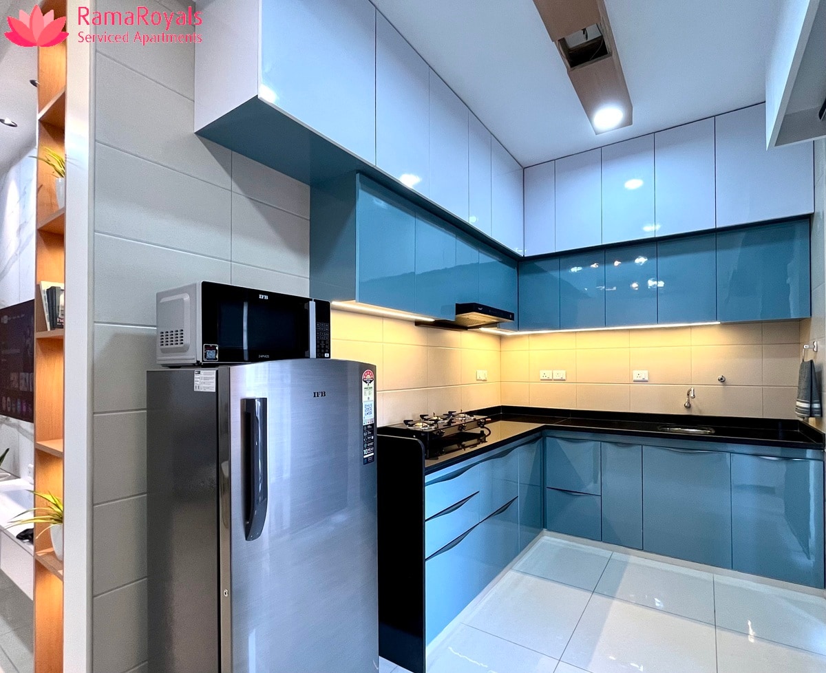 The kitchen features modern cabinetry with a blue and white color scheme. A stainless steel refrigerator, microwave, and gas stove are visible. Under-cabinet lighting enhances the workspace, while tiles on the floor reflect the sleek design.