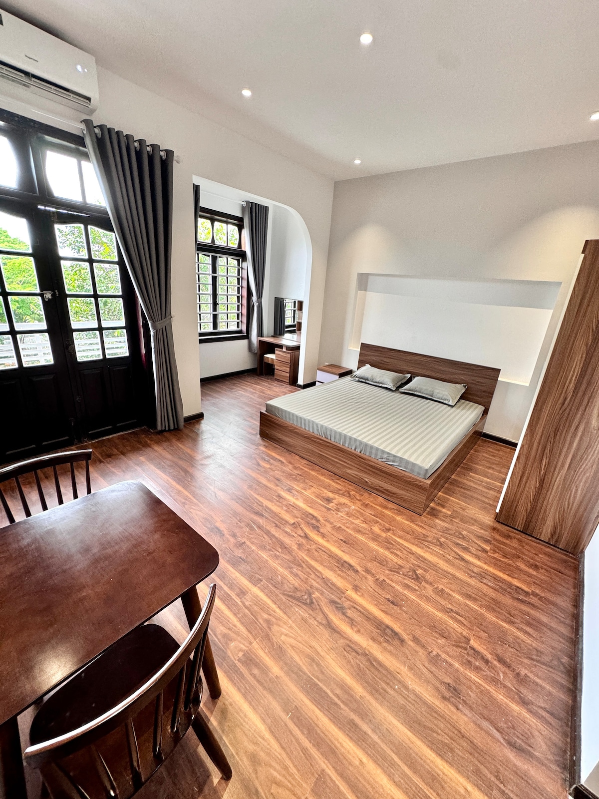 An airy room features a spacious bed with light gray bedding. A wooden table and two chairs are positioned nearby. Large windows with dark curtains allow natural light to enter, while wooden flooring adds warmth to the area.