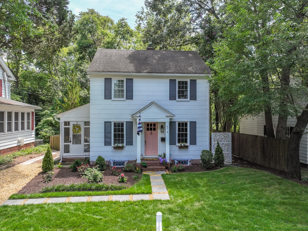 The exterior of a charming two-story home is framed by well-maintained greenery and colorful flower beds. A welcoming front porch with a pink door contributes to the overall inviting appearance. A neatly arranged pathway leads to the entrance, bordered by manicured lawns.