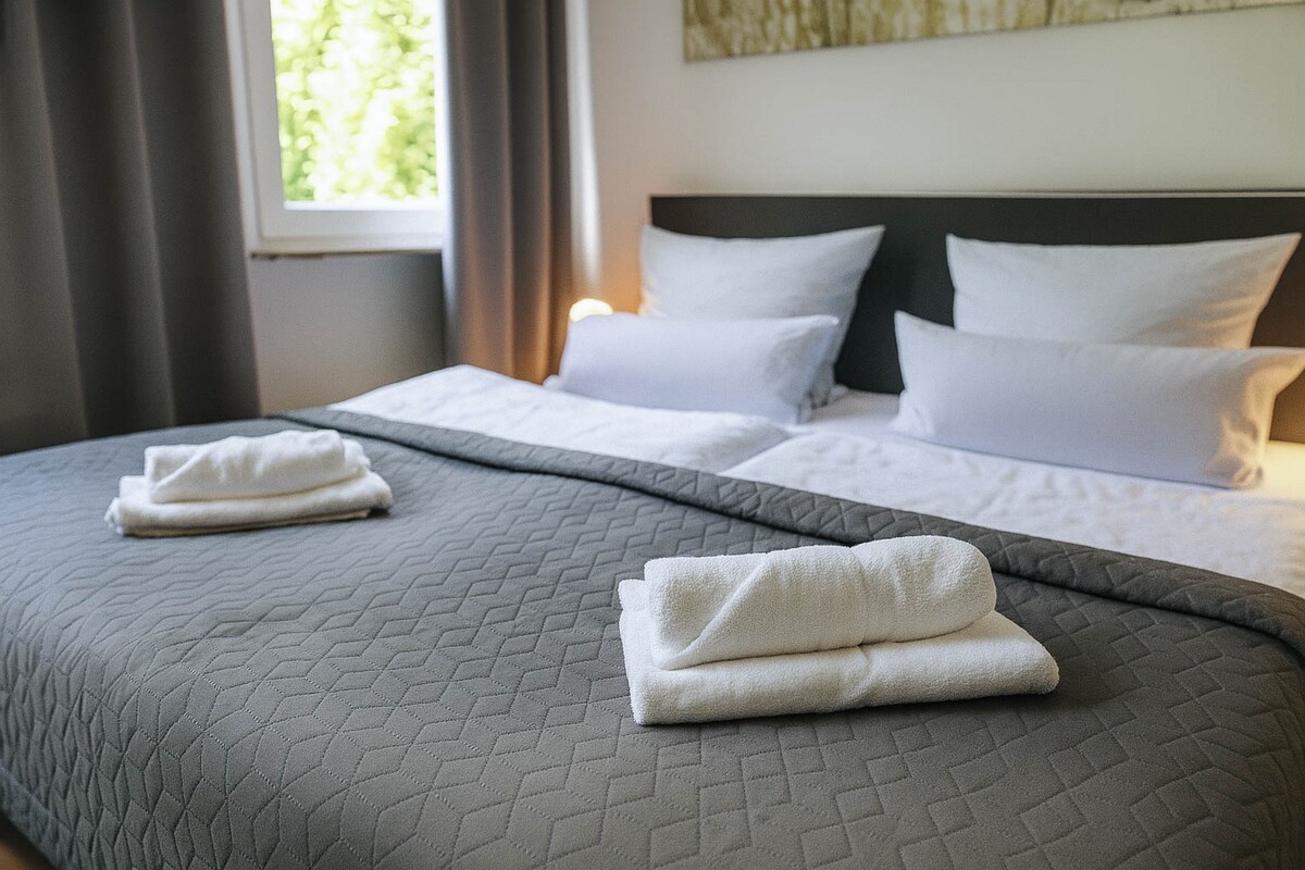 A spacious bed is dressed in light bedding, featuring white pillows and a grey quilted blanket. Two neatly folded white towels are placed at the foot of the bed. Natural light filters in through a nearby window, complemented by grey curtains.