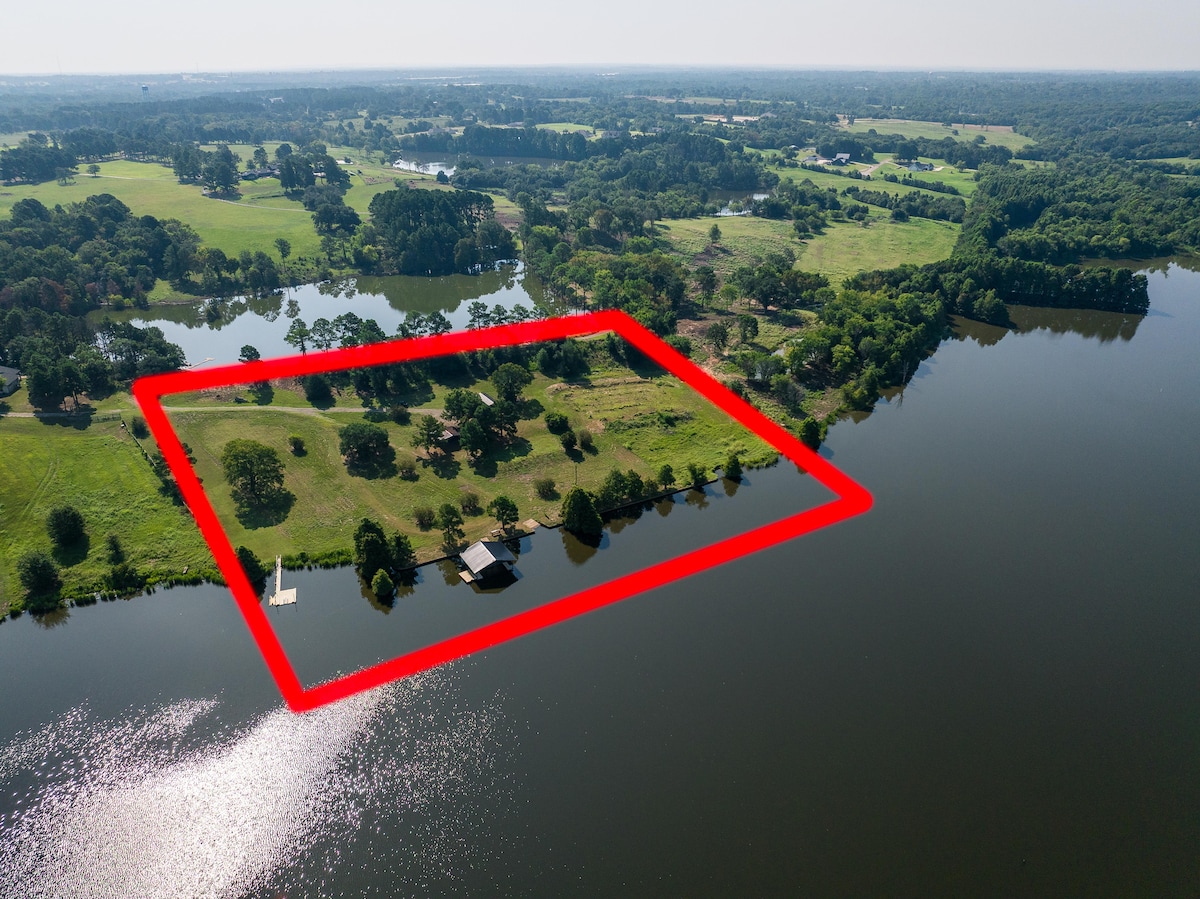 An aerial view of a spacious property bordered by lush greenery and the calm waters of a private lake. The rectangle highlights the extensive area, showcasing well-maintained grass and trees. A cabin and boat dock are visible along the shoreline, offering a serene natural landscape.