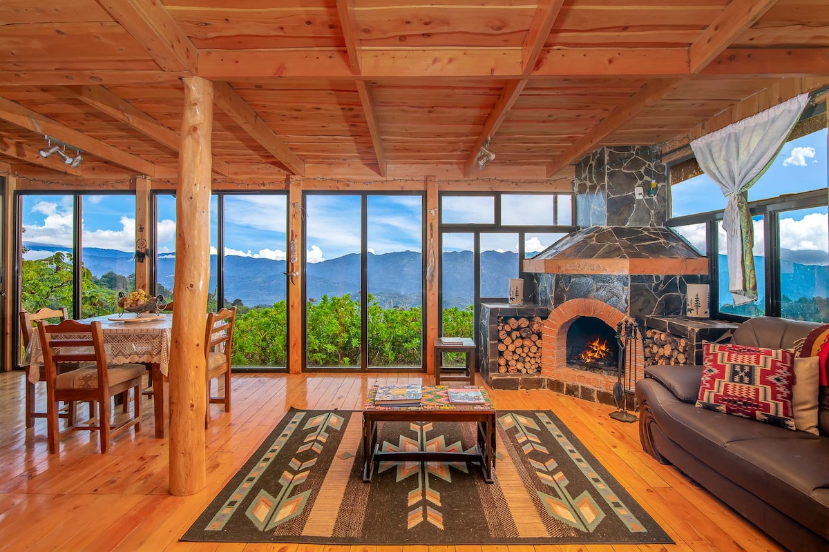 The living area features large glass windows that frame expansive mountain views. A wood-burning fireplace is surrounded by rustic stone, with logs neatly stacked nearby. Comfortable seating and a wooden dining table enhance the cozy atmosphere, complemented by the warm wood ceiling and flooring.